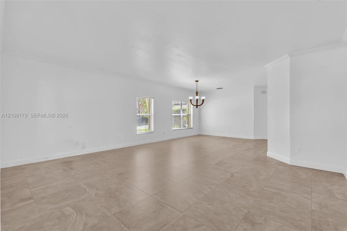 Open-concept interior featuring large-format tile flooring, recessed crown molding, dual windows, and a multi-arm chandelier