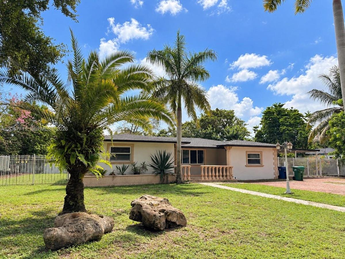 Single-story residence featuring a light-colored stucco exterior, decorative stone balustrade, mature palm trees, manicured lawn, and perimeter metal 