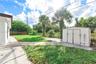 Rear exterior featuring a concrete patio, freestanding storage shed, open lawn space, perimeter fencing, and mature palm trees