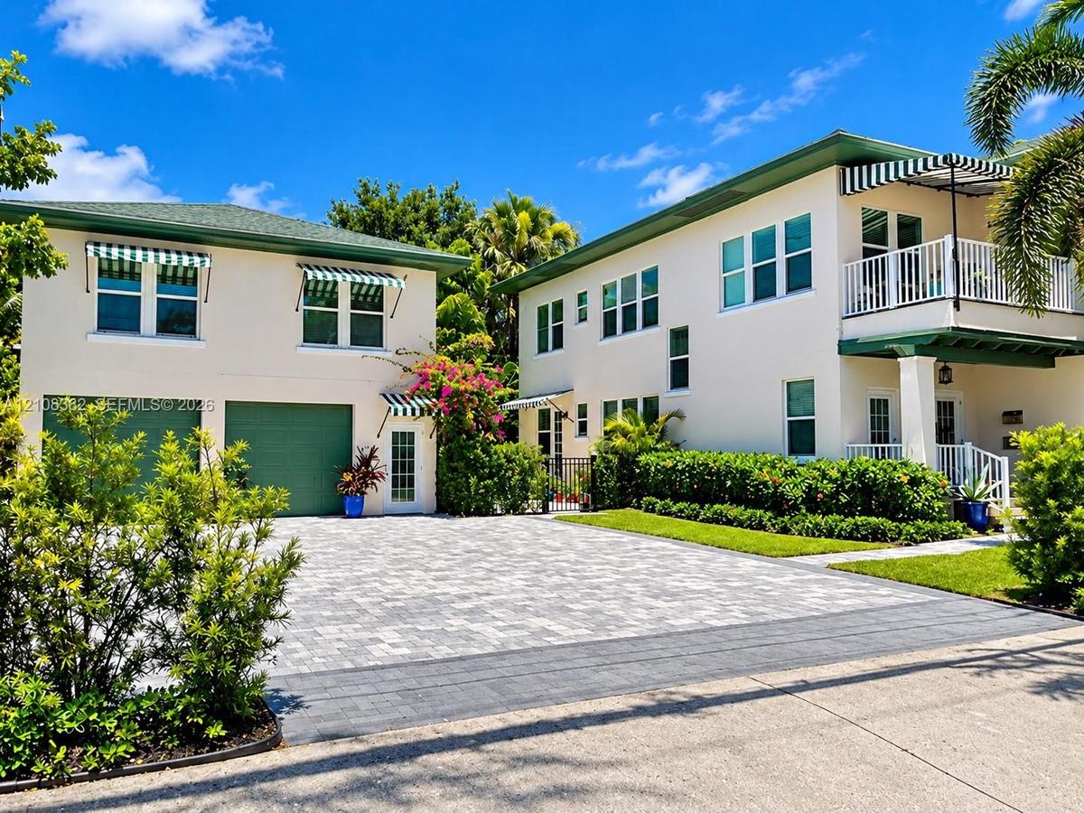 Two-story exterior featuring multi-tone paver driveway, green-tinted garage doors, striped fabric awnings, a second-story balcony with white railing, 