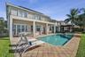 Stucco exterior featuring a second-story balcony with metal railings, a covered ground-level patio, and a concrete-paver pool deck surrounding an ingr