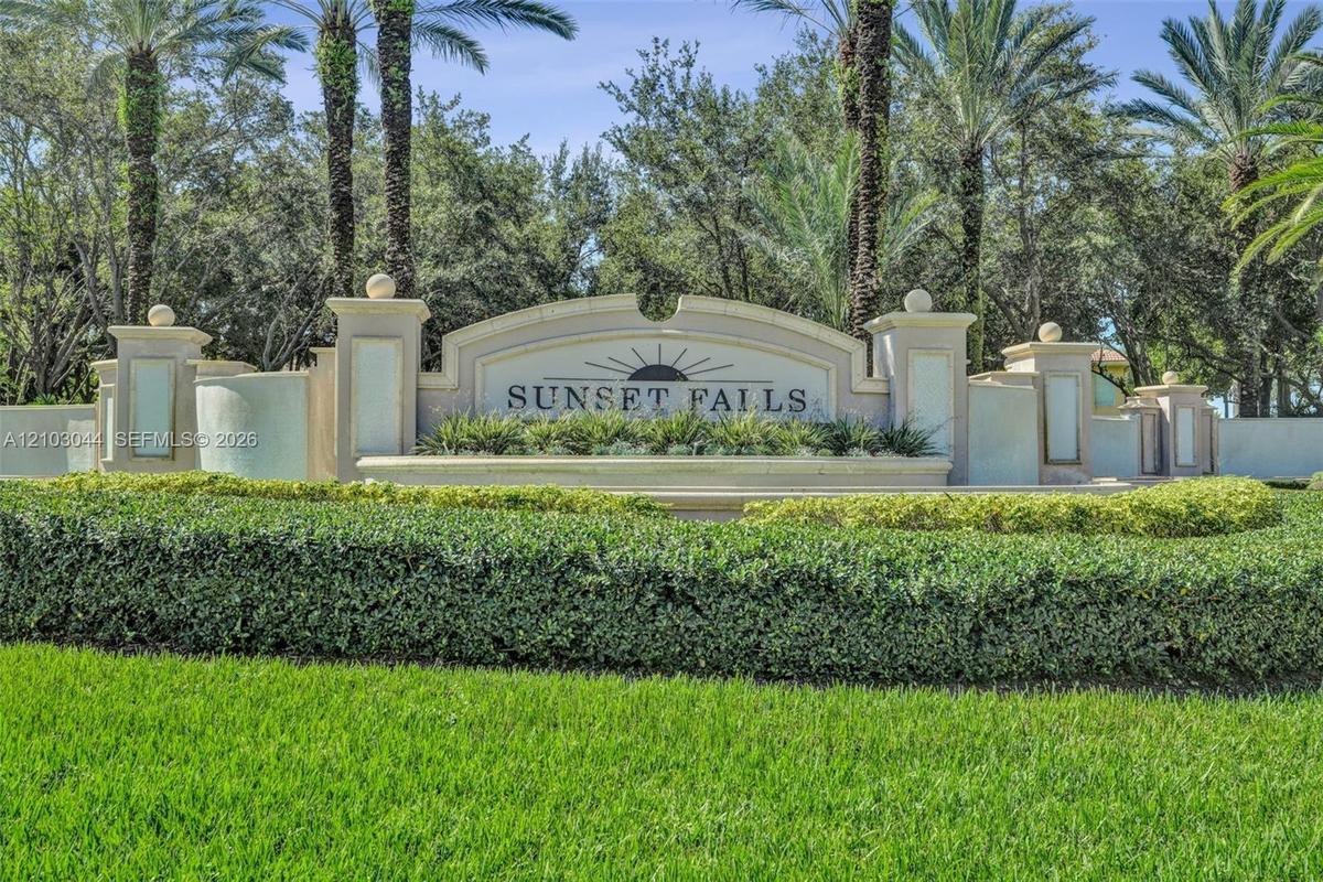 Stucco community signage featuring decorative masonry pillars, integrated lighting fixtures, tiered architectural detailing, and mature palm landscapi