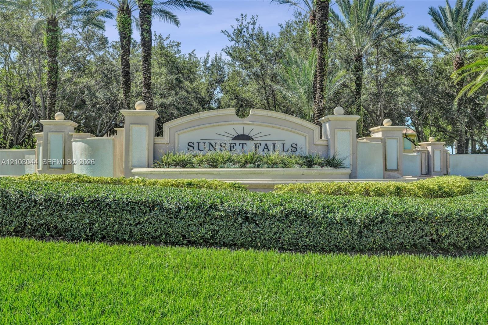 Stucco community signage featuring decorative masonry pillars, integrated lighting fixtures, tiered architectural detailing, and mature palm landscapi