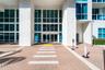 Grand entrance featuring multi-story floor-to-ceiling glass paneling, white structural pillars, and a herringbone-patterned brick paver arrival area