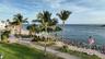 Waterfront property featuring a paved promenade, mature palm trees, expansive lawn, and a rock-lined shoreline