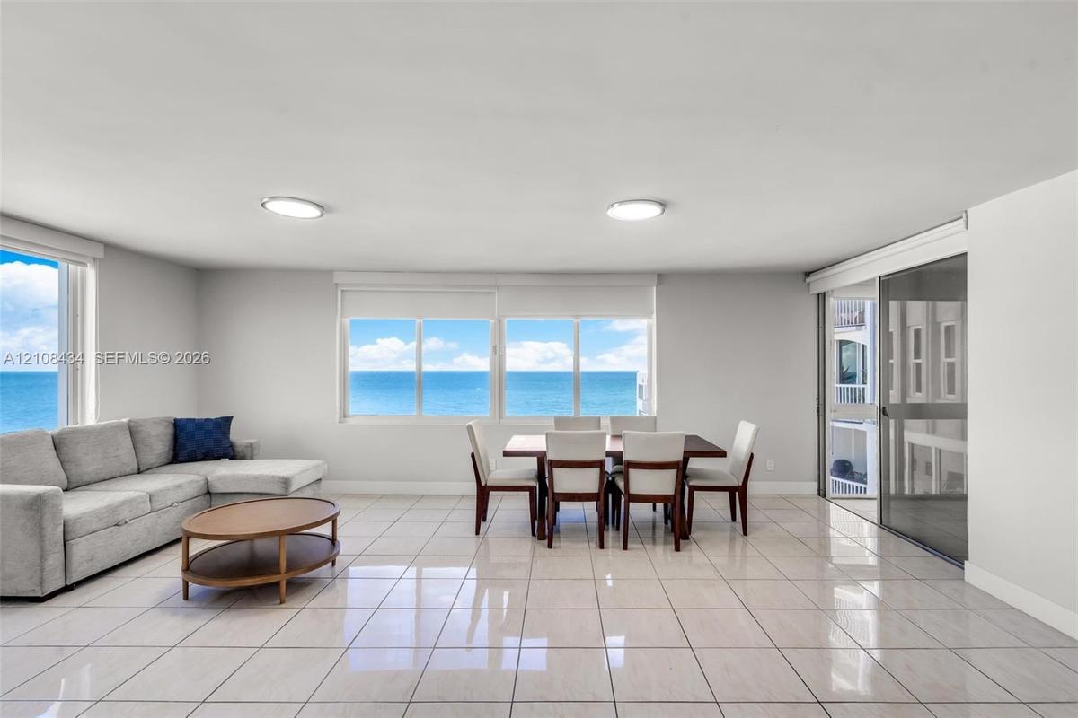 Open-concept room featuring light-toned tile flooring, large multi-pane windows, and a sliding glass door providing access to a balcony with an ocean 
