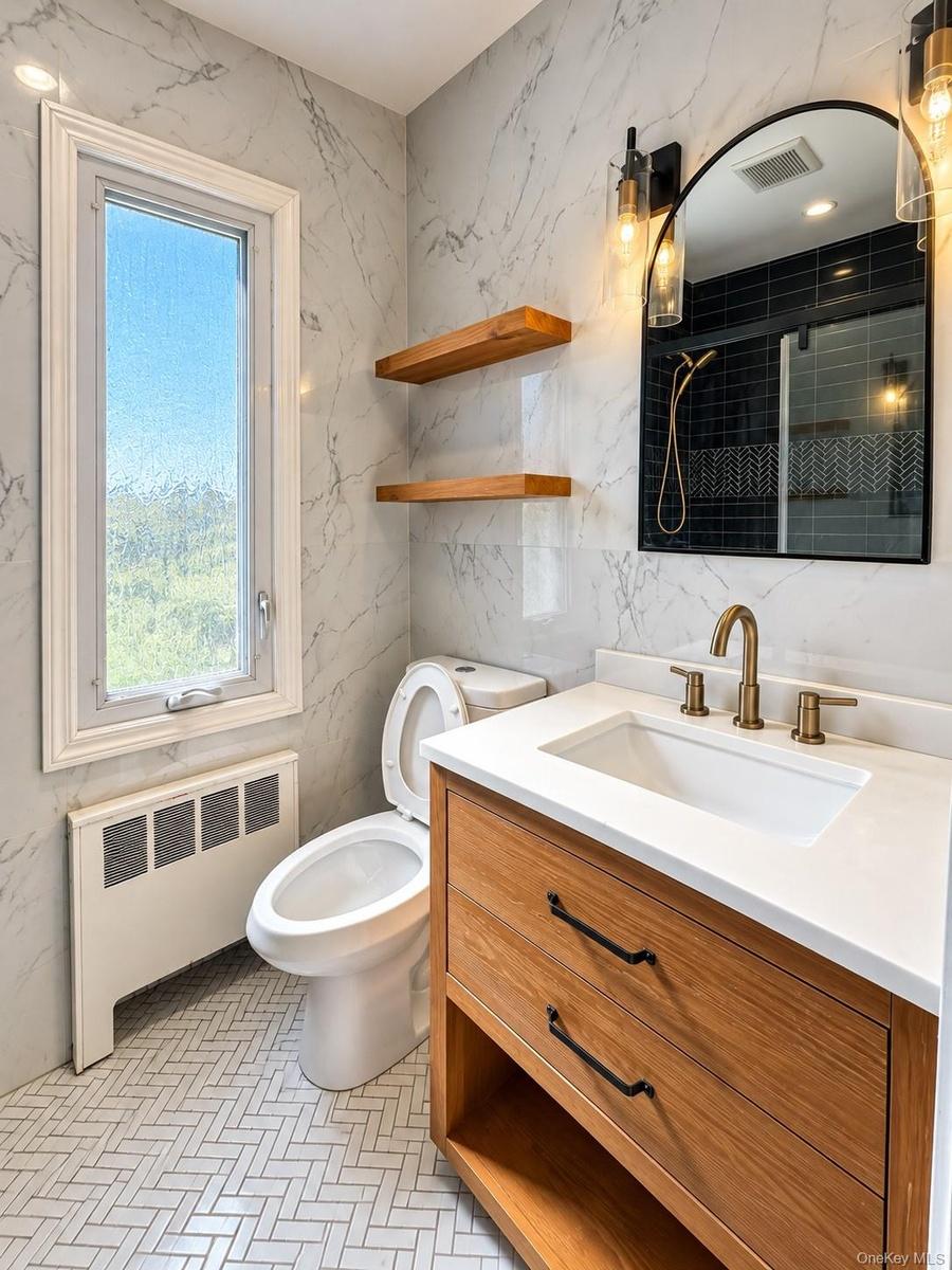 Bathroom featuring stone-finish wall tile, custom wood-finish vanity with a stone countertop, arched wall-mounted mirror, floating wood shelving, and 