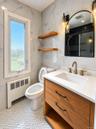 Bathroom featuring stone-finish wall tile, custom wood-finish vanity with a stone countertop, arched wall-mounted mirror, floating wood shelving, and 