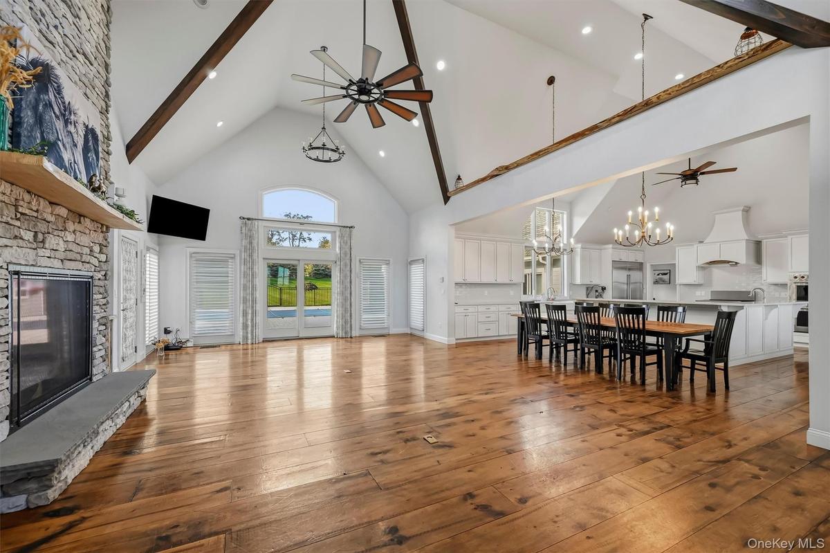 Vaulted ceilings with exposed timber beams, floor-to-ceiling stone fireplace, wide-plank wood-finish flooring, and natural light via arched glass door