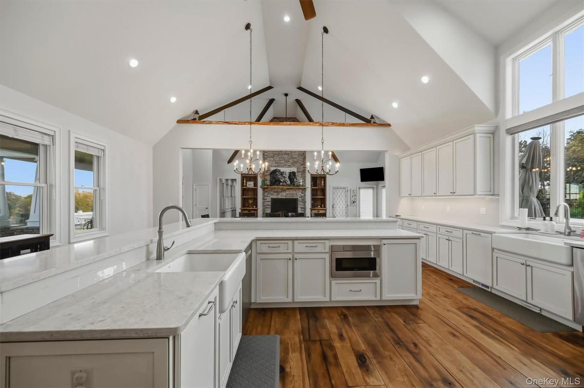 Vaulted ceiling with exposed timber beams, white shaker cabinetry, quartz surfaced countertops, apron-front farmhouse sinks, and wood-finish flooring