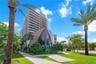 Multi-story architectural exterior featuring a curved metallic-toned facade, floor-to-ceiling glass paneling, tiered balconies, and manicured grounds 