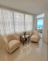Floor-to-ceiling pleated window treatments, polished tile flooring, and a glass-paneled door leading to an outdoor terrace with ocean scenery