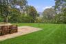 Expansive lawn featuring a concrete patio, timber retaining wall, mature perimeter trees, and a chain-link fence