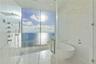 Floor-to-ceiling glass paneling, freestanding soaking tub, walk-in shower with multi-head wall-mounted shower system, and light-toned tile flooring