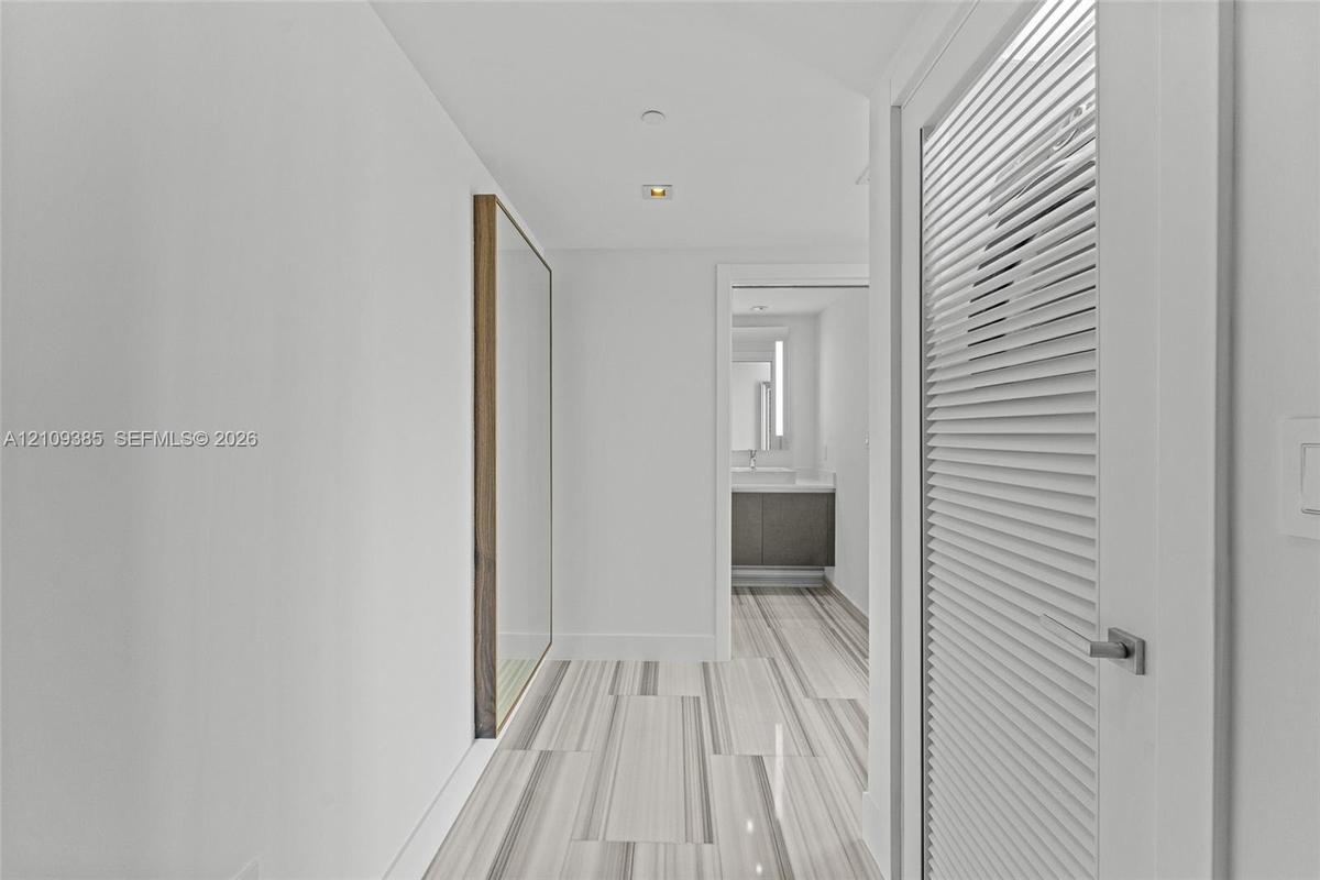 Hallway featuring linear-patterned tile flooring, a louvered white door with satin hardware, recessed ceiling lighting, and a flush-mounted wall mirro