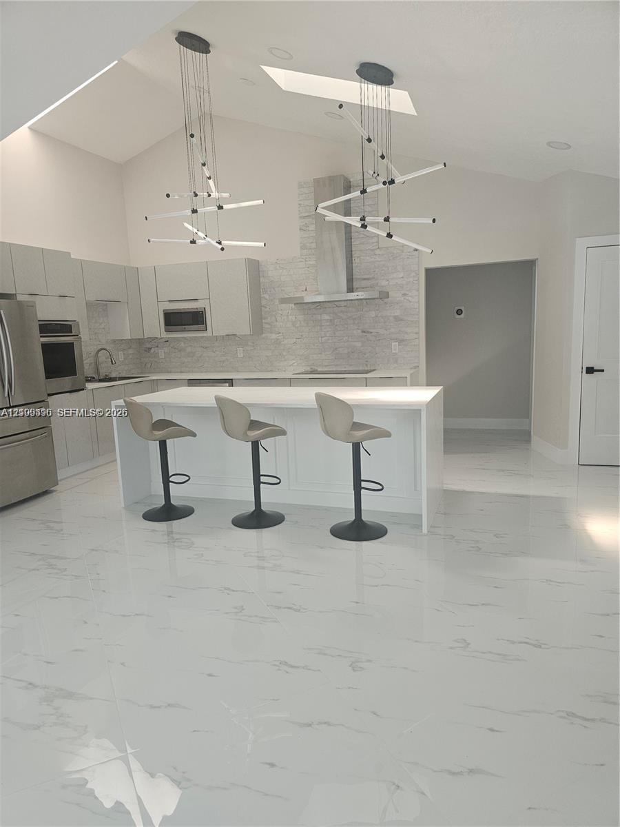Kitchen featuring vaulted ceilings, skylights, white cabinetry, a center island with solid surfacing, and a stone-tiled backsplash with a stainless st