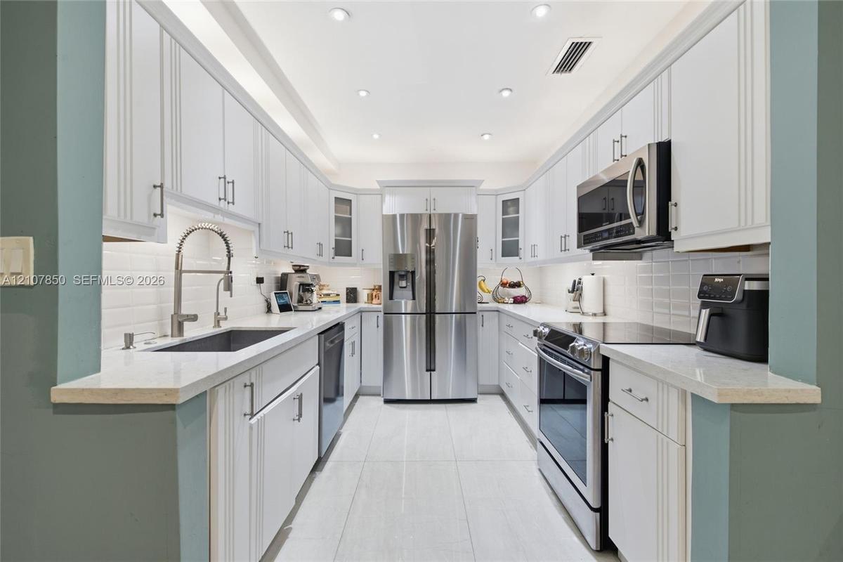Galley kitchen featuring white cabinetry, light-toned stone-finish countertops, stainless steel appliances, a white tile backsplash, and recessed ligh