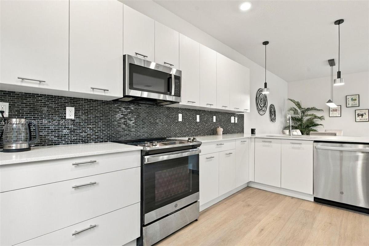 Kitchen featuring white flat-panel cabinetry, light-toned stone countertops, black mosaic tile backsplash, stainless steel appliances, and wood-finish