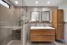 Floating wood-finish double vanity with integrated sinks, glass-enclosed walk-in shower with mosaic tile accents, matching wood-finish tall cabinet, a