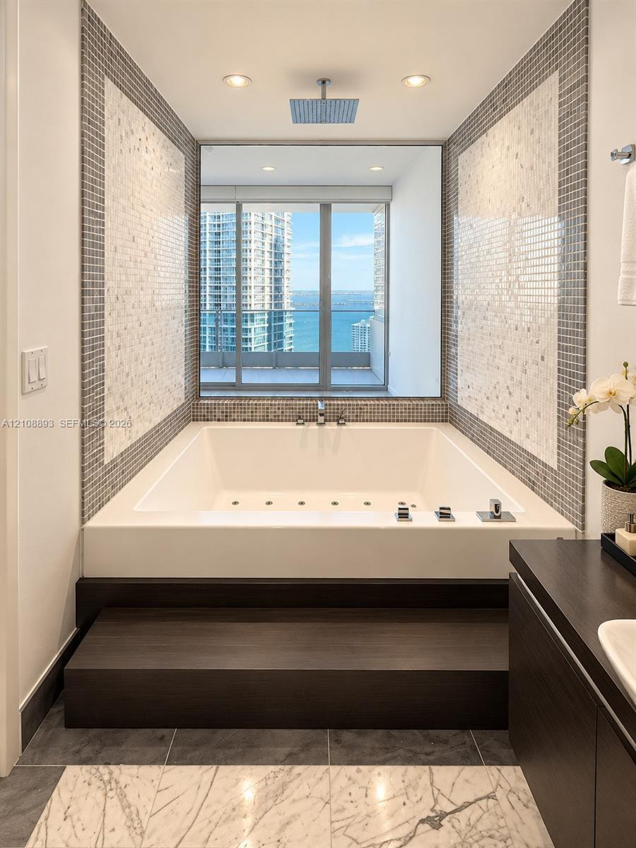 Built-in soaking tub with a raised dark wood-finish platform, overhead rainfall showerhead, mosaic tile surround, and expansive window showcasing wate