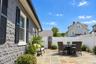 Exterior featuring grey rock-faced masonry siding, dark window shutters, natural stone paver patio, white vinyl privacy fencing, and metal decorative 