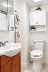 Wall-mounted white cabinetry with open shelving, wood-finish vanity, polished chrome faucet, rectangular wall-mounted mirror, and standard-height toil
