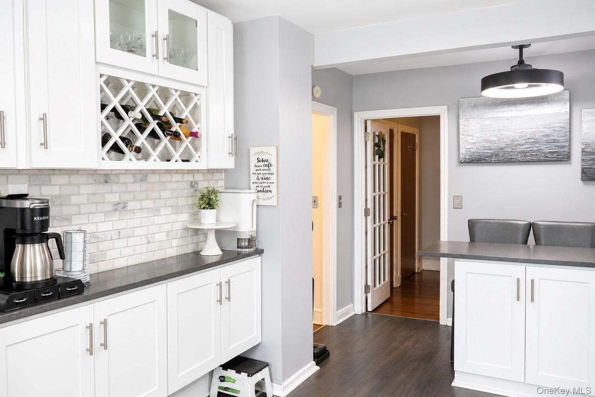 Kitchen featuring white cabinetry with integrated wine rack, stone-tiled backsplash, dark solid-surface countertops, wood-finish flooring, and a moder