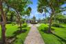 Expansive grounds featuring a brick paver walkway, manicured lawn, established shade trees, and mature tropical landscaping