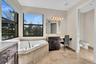 Corner soaking tub with mosaic tile surround, large windows, dark wood cabinetry with light countertops, and neutral tile flooring