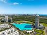 Aerial perspective of a high-rise residential tower featuring a large man-made lagoon with a sandy shoreline, surrounding parkland, and coastal city s