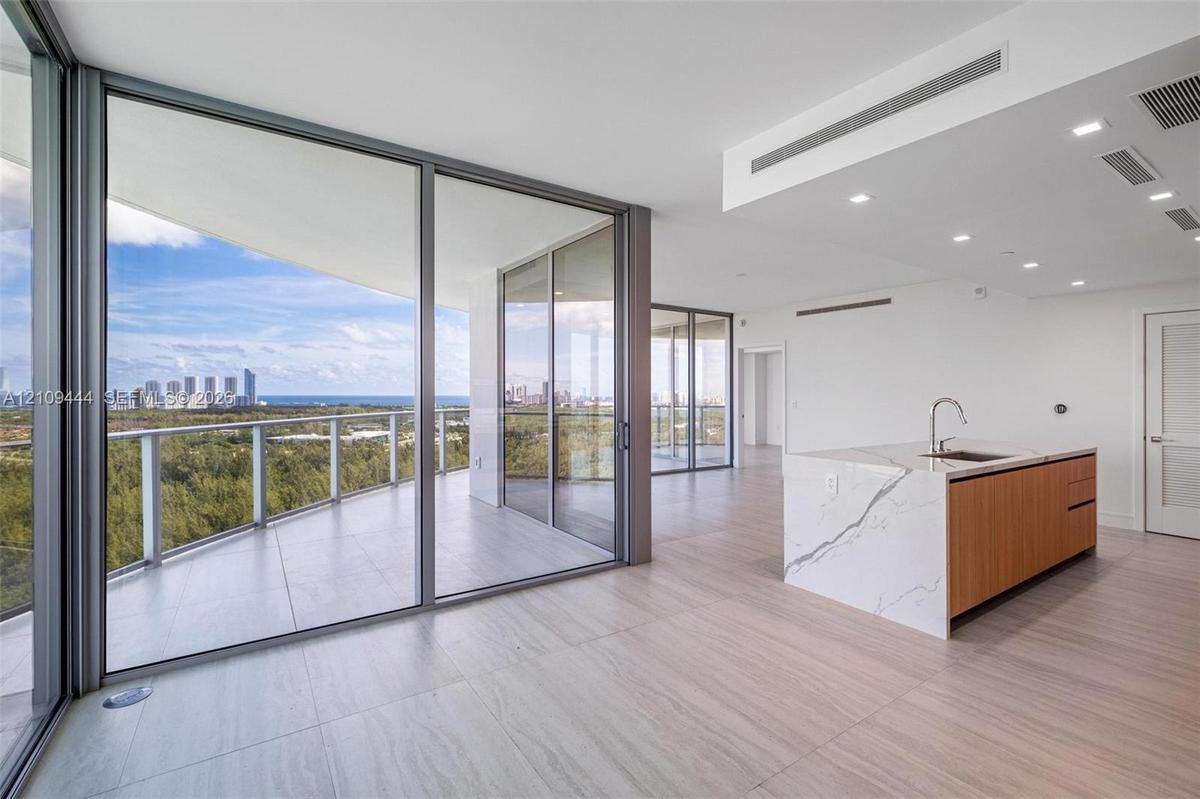Floor-to-ceiling glass walls, expansive wraparound balcony, kitchen island with waterfall-edge stone countertop, wood-finish cabinetry, and neutral la
