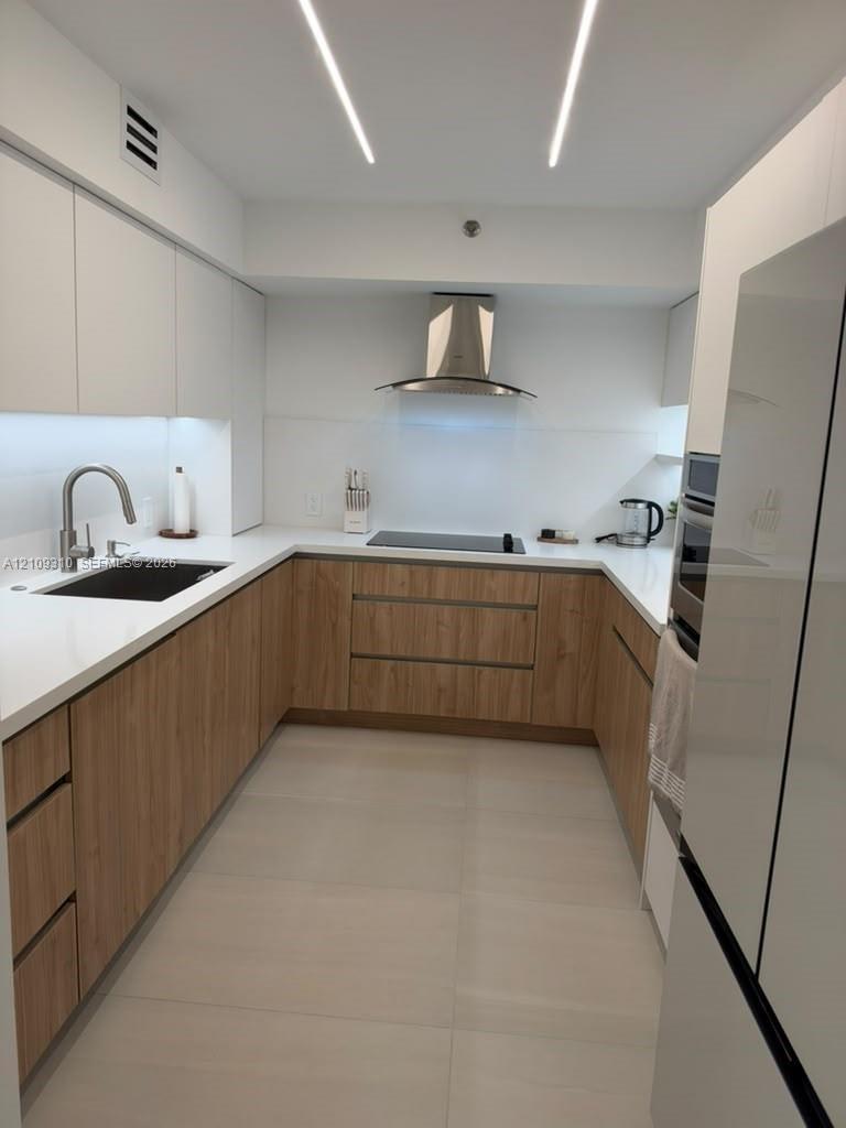 Modern kitchen featuring wood-finish cabinetry, solid-surface countertops, undermount sink with gooseneck faucet, stainless steel range hood, and line