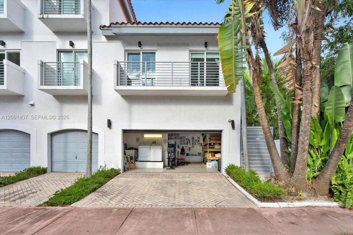 Stucco exterior featuring dual-level metal balconies, terracotta-style roof tile accents, a private herringbone-patterned paver driveway, and an attac