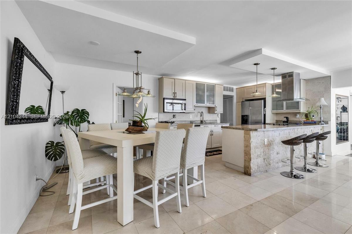 Open-concept kitchen featuring light wood cabinetry, stone-clad kitchen island, stainless steel appliances, tiered tray ceilings, and neutral tile flo
