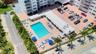 Aerial view of the building exterior featuring an in-ground swimming pool, paved pool deck, multi-level parking area, and landscaping with palm trees