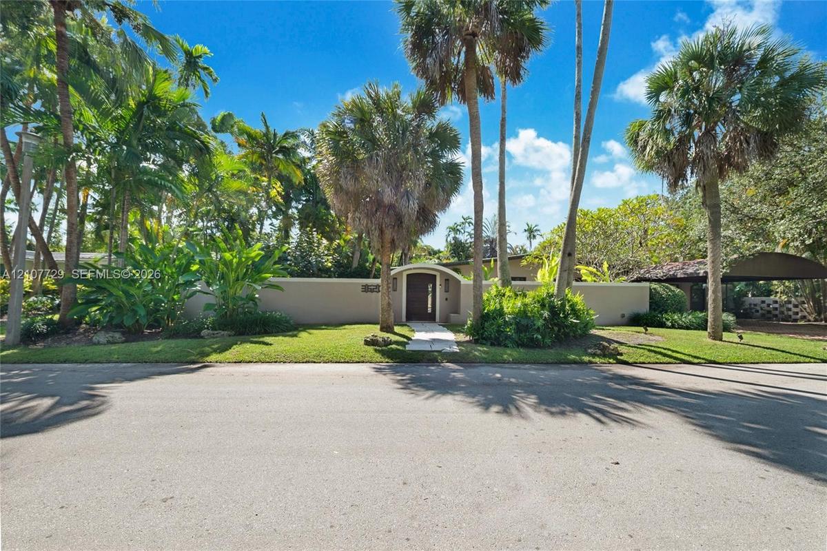 Exterior property featuring a privacy wall, arched entryway with a solid door, paved walkway, mature palms, and a covered carport