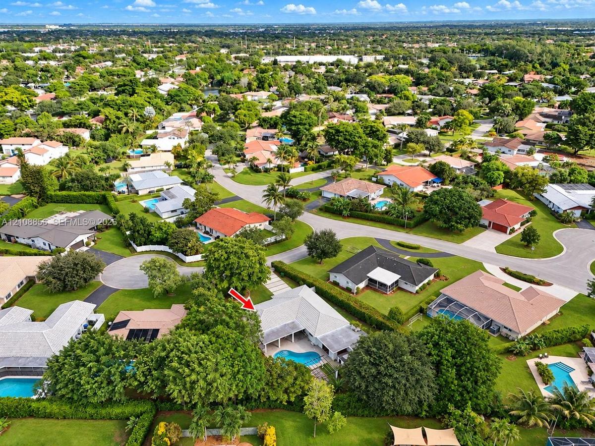 Aerial perspective of a residential property featuring an in-ground swimming pool, covered patio, concrete driveway, and mature landscaping