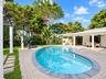 Kidney-shaped swimming pool with stone coping, expansive paver patio, covered porch with classical columns, and mature landscaping