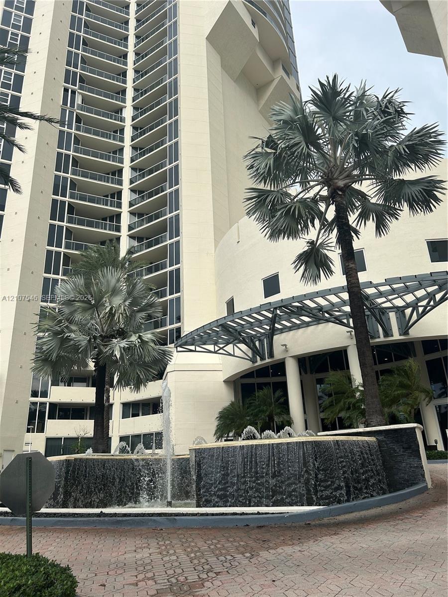 High-rise exterior featuring curved glass balconies, a circular fountain with tiered stone masonry, a metal-framed porte-cochere, and mature palm land