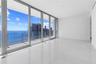 Floor-to-ceiling glass panel windows, wide-plank tile flooring, and a glass-railed exterior balcony featuring a water and cityscape backdrop