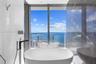 Freestanding soaking tub featuring chrome floor-mounted faucet, floor-to-ceiling windows with water backdrop, and large-format tile wall surrounds