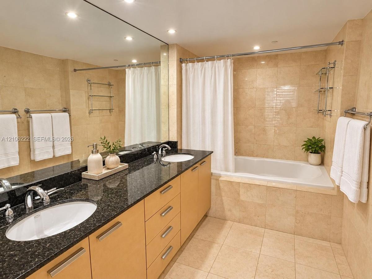 Double vanity with dark stone countertop, integrated dual sinks, light wood-finish cabinetry, floor-to-ceiling tile work, and built-in soaking tub