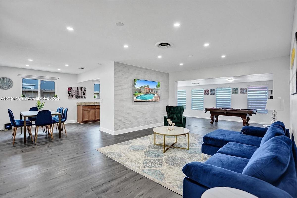 Open-concept interior featuring wood-finish flooring, recessed lighting, a painted brick accent wall, and horizontal window blinds