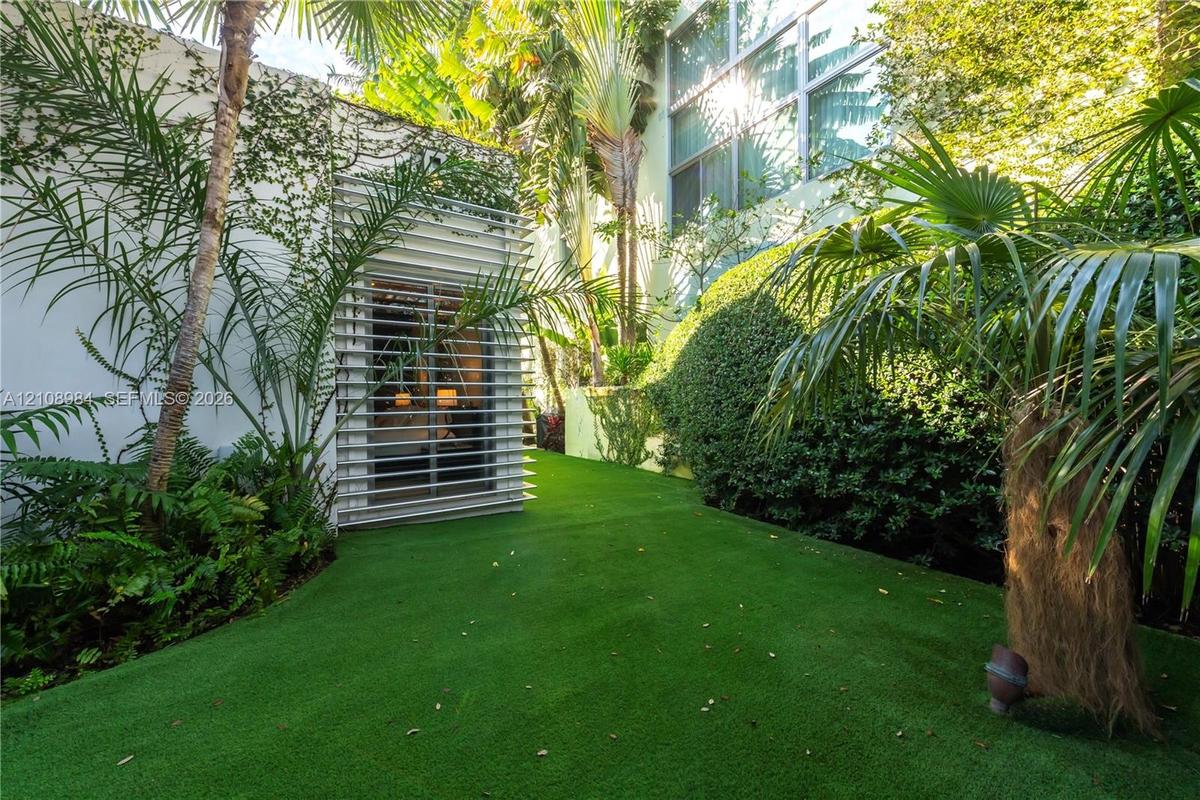 Private exterior space featuring synthetic turf, structured louvered window detailing, white stucco wall surfaces, mature tropical landscaping, and la