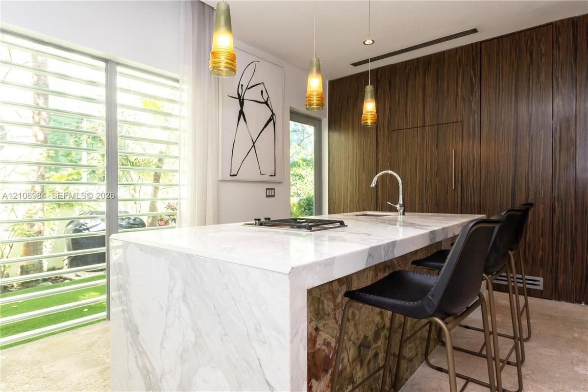 Waterfall-edge stone island with integrated cooktop and undermount sink, floor-to-ceiling wood-finish cabinetry, triple glass pendant lighting, and fu