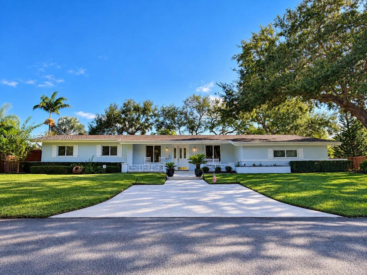 Single-story residence featuring light-toned exterior cladding, a paved walkway, covered front porch with decorative railing, and manicured landscapin
