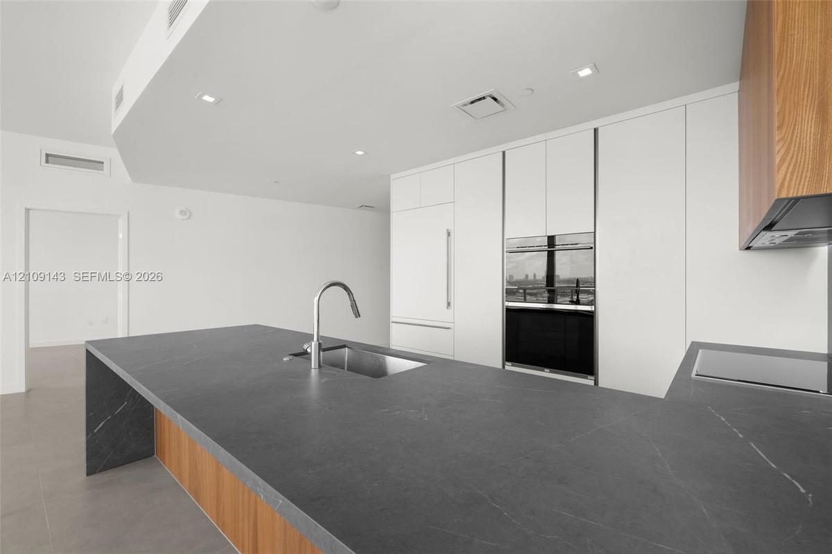 Kitchen featuring a waterfall-edge island with dark stone countertop, undermount stainless steel sink, flat-panel white cabinetry, wood-finish lower c