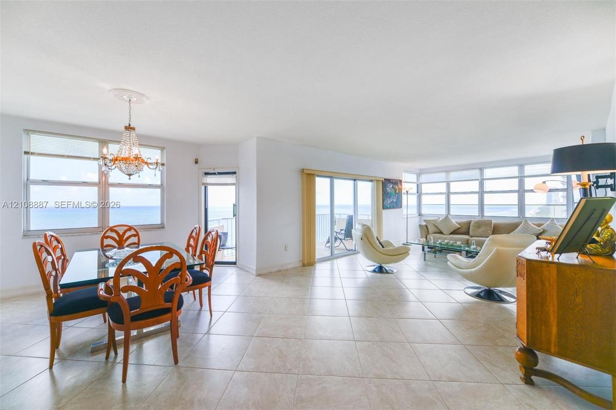 Open-concept main living area featuring light-toned tile flooring, oversized windows, multi-pane sliding glass doors, and a decorative crystal chandel