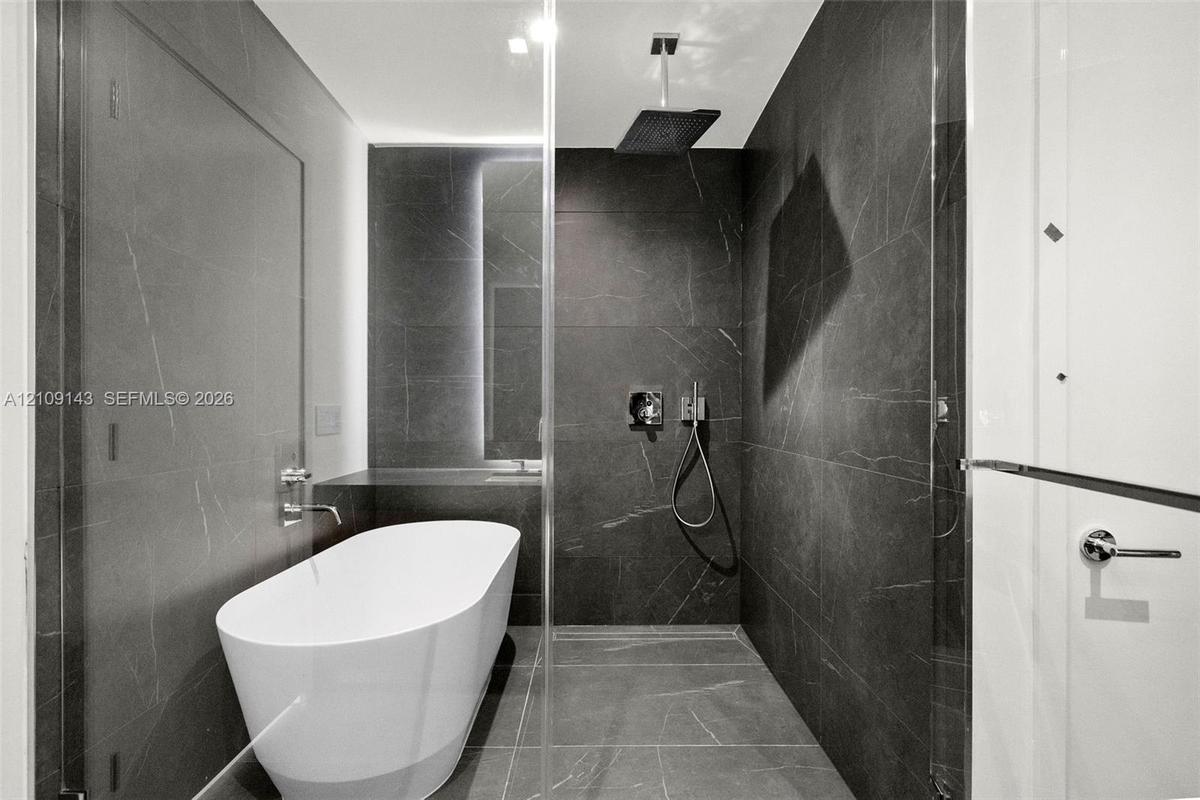 Freestanding soaking tub, large-format dark tile wall surfacing, overhead rainfall showerhead, handheld shower fixture, and frameless glass enclosure