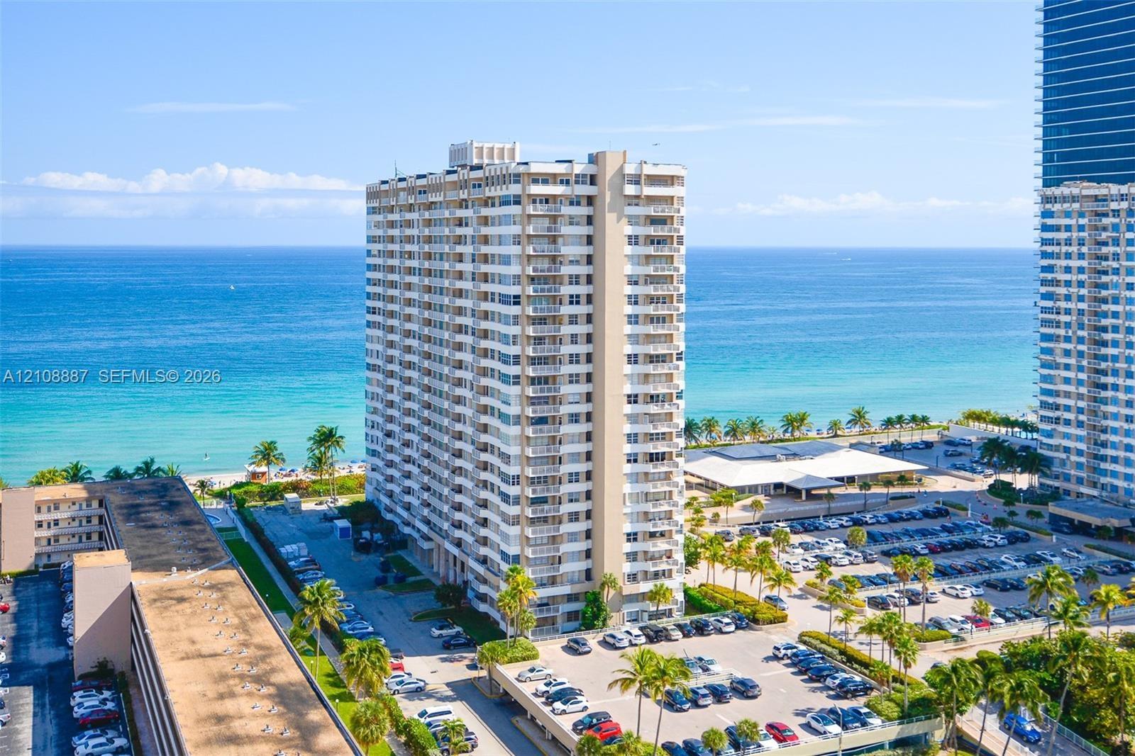 High-rise residential building featuring stacked private balconies, an expansive paved parking area, and mature palm tree landscaping